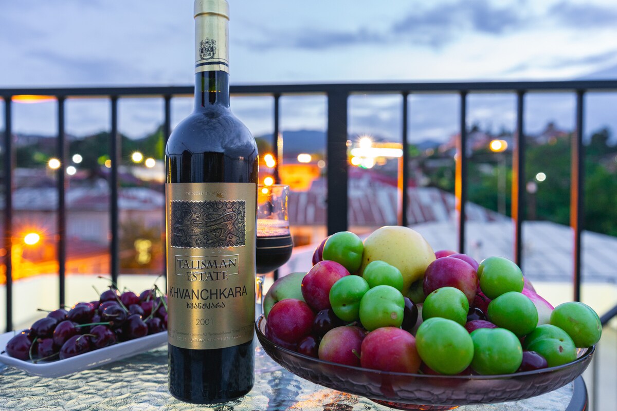 A bottle of Khvanchkara wine is placed beside a glass filled with red wine. A bowl of assorted fruits, including green and red apples, cherries, and plums, is displayed on a reflective surface, with the city lights and sky visible in the background.