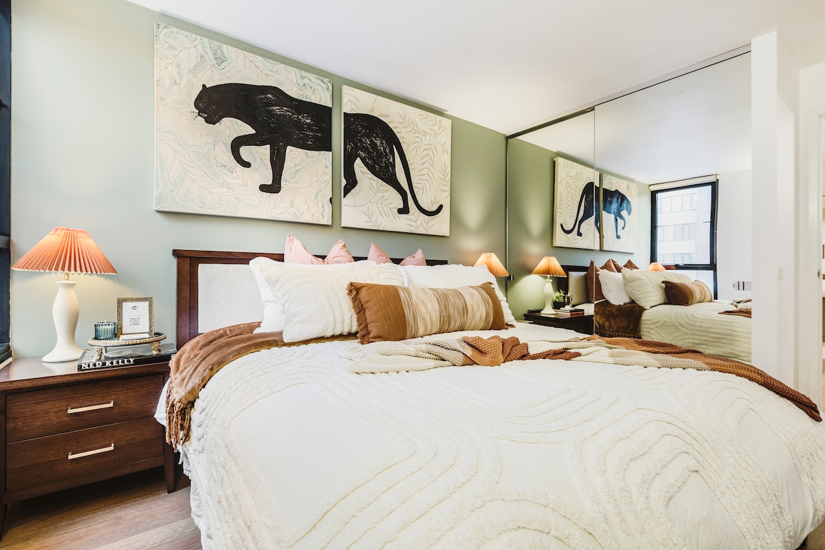 A spacious bedroom features a king size bed covered with a white quilt and accented by warm-toned pillows. Two bedside tables are adorned with lamps, while large artistic wall hangings of a panther add character. A mirrored wardrobe reflects the room's design.