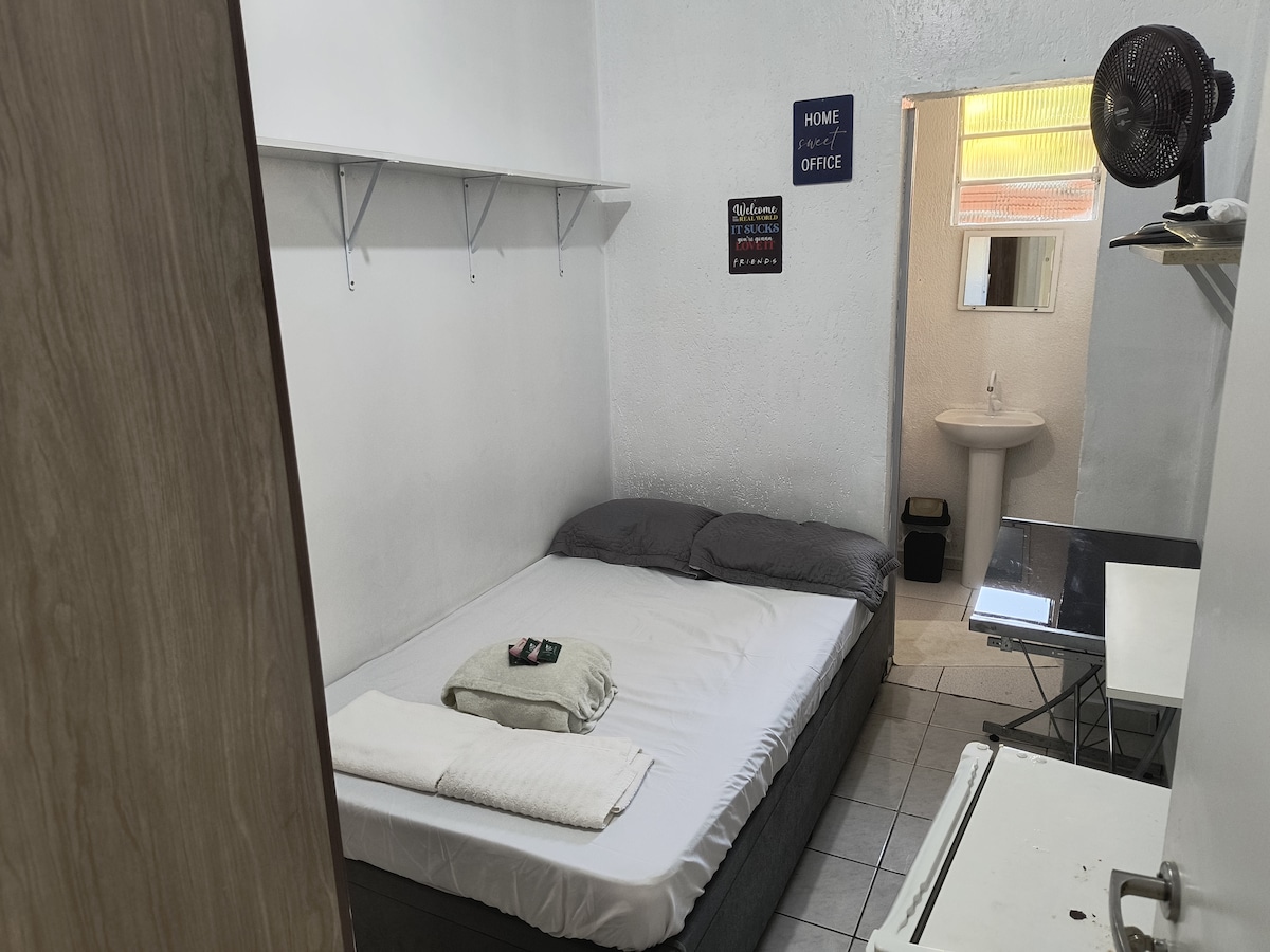A cozy bedroom features a spacious bed with neutral linens and two pillows. A small table, fan, and shelves are visible against the walls. A sink and mirror are present in the adjoining area, enhancing the practicality of the space.