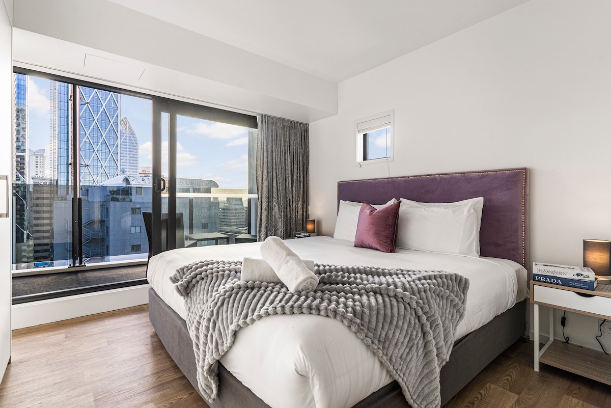 A spacious bedroom features a super king-size bed adorned with a textured grey throw. Large windows provide ample natural light, showcasing city views. Soft lighting from bedside lamps enhances the serene ambiance. A cozy decorative pillow adds a touch of comfort to the bedding arrangement.