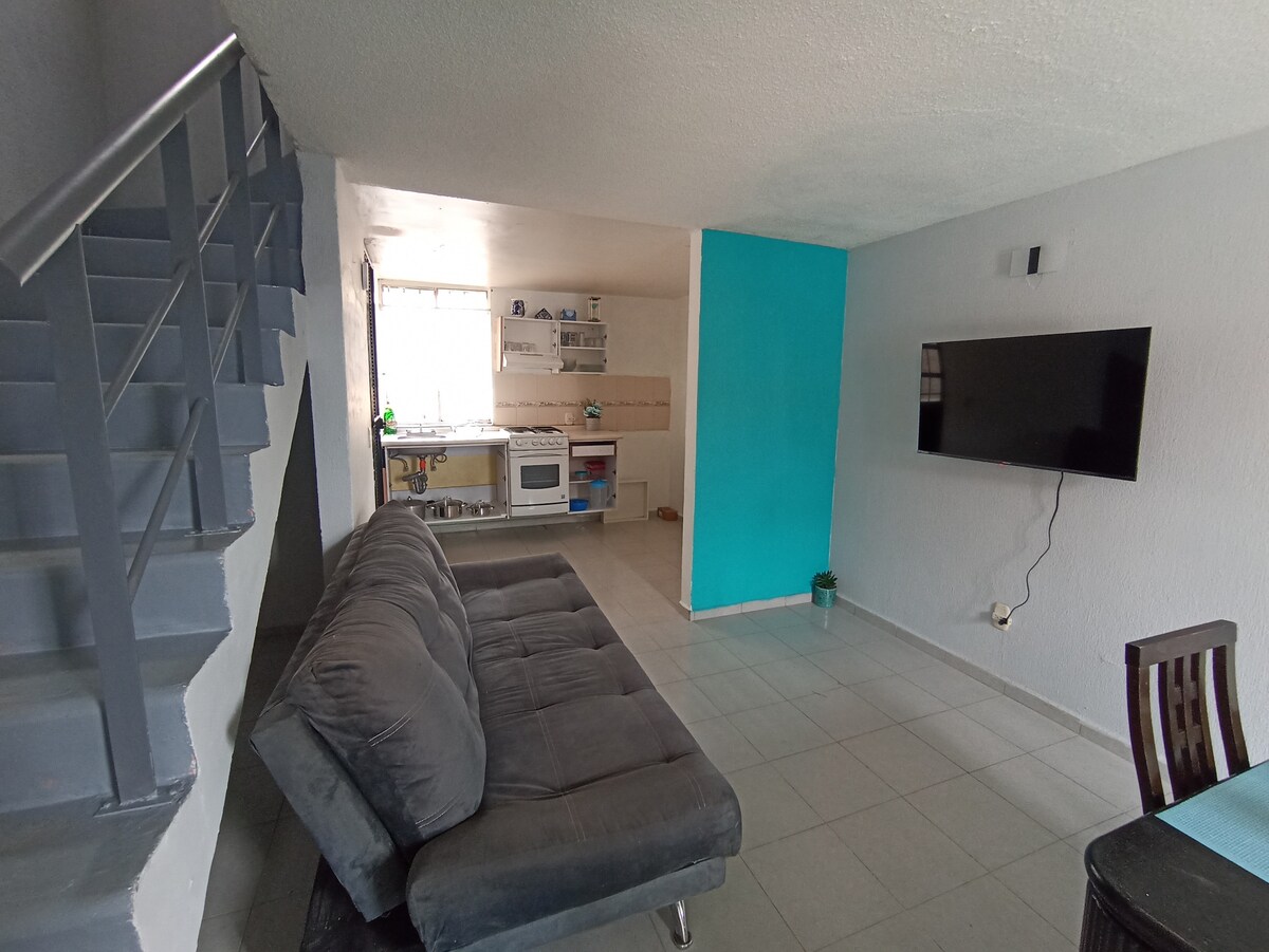 The living area features a spacious gray sofa and a wall-mounted television. A bright blue accent wall contrasts with the neutral tones. A staircase is present on the left side, and a view of a well-equipped kitchen with natural light can be seen in the background.