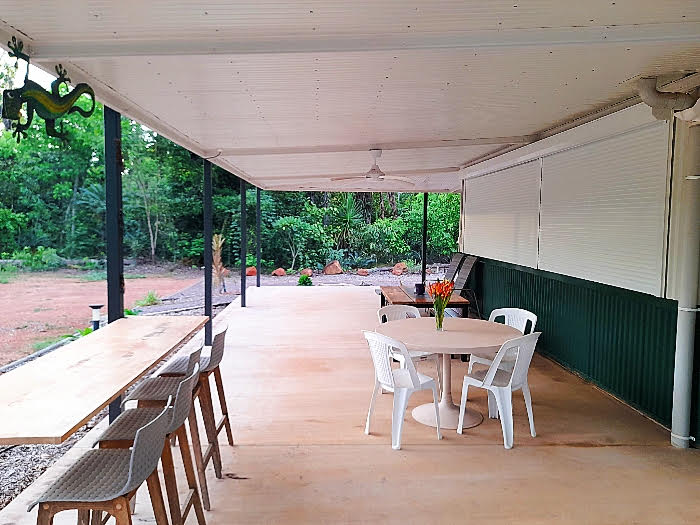 An expansive outdoor entertainment area features a large concrete floor covered by a roof. A round dining table with chairs and two long tables for seating are visible, surrounded by lush greenery. Ceiling fans and a decorative element add to the relaxed atmosphere.