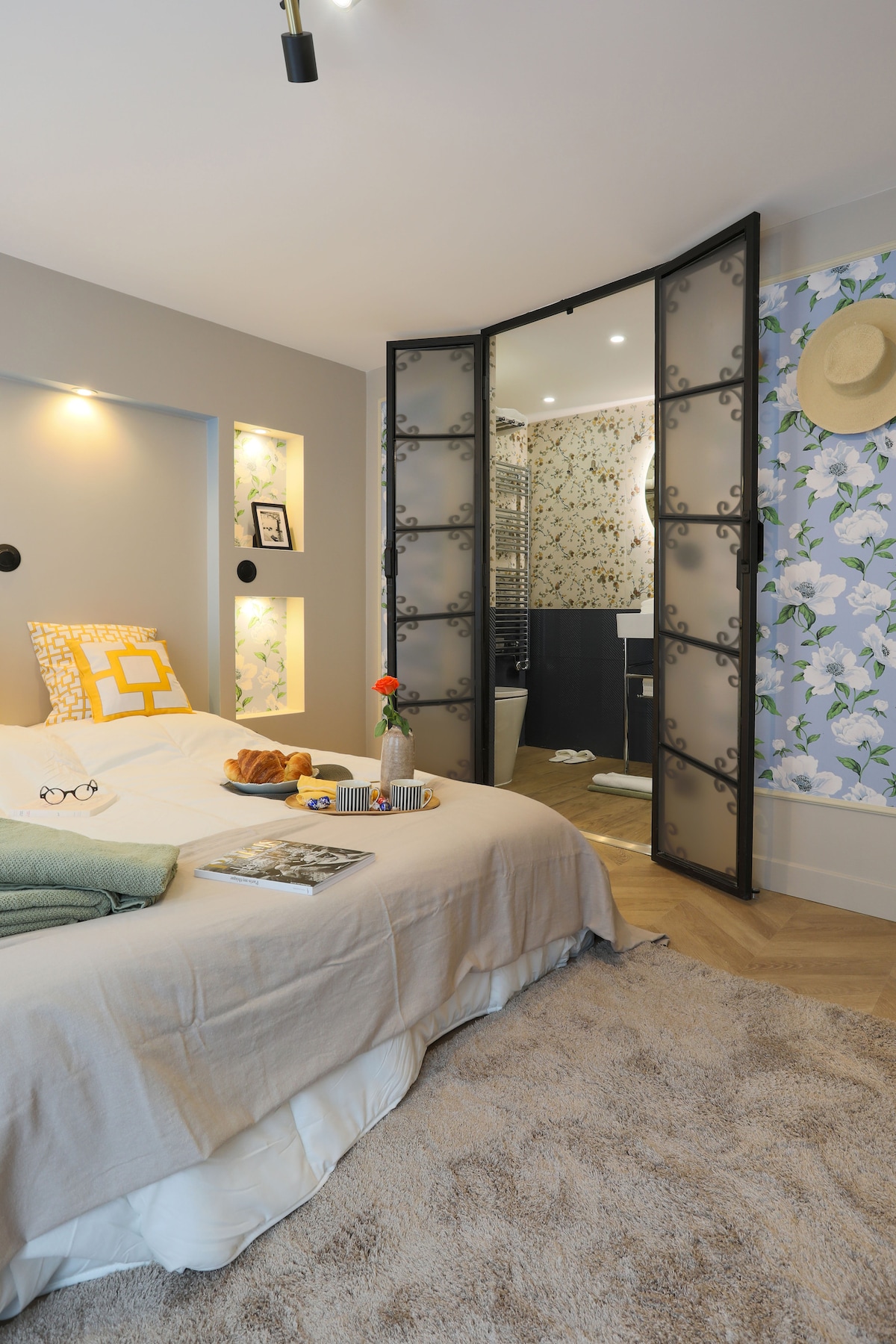 A comfortable sleeping area is presented with a spacious bed adorned with soft linens and decorative pillows. A cozy rug covers the floor, and a small vase of flowers adds a touch of warmth. A partition with a decorative design leads to the modern bathroom.