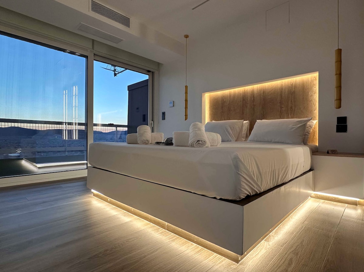 A bright bedroom with a spacious bed featuring soft linens and stylish pillows. Ambient lighting outlines the bed's base, adding a subtle glow. Floor-to-ceiling windows offer expansive views of the cityscape, while modern hanging light fixtures enhance the design.