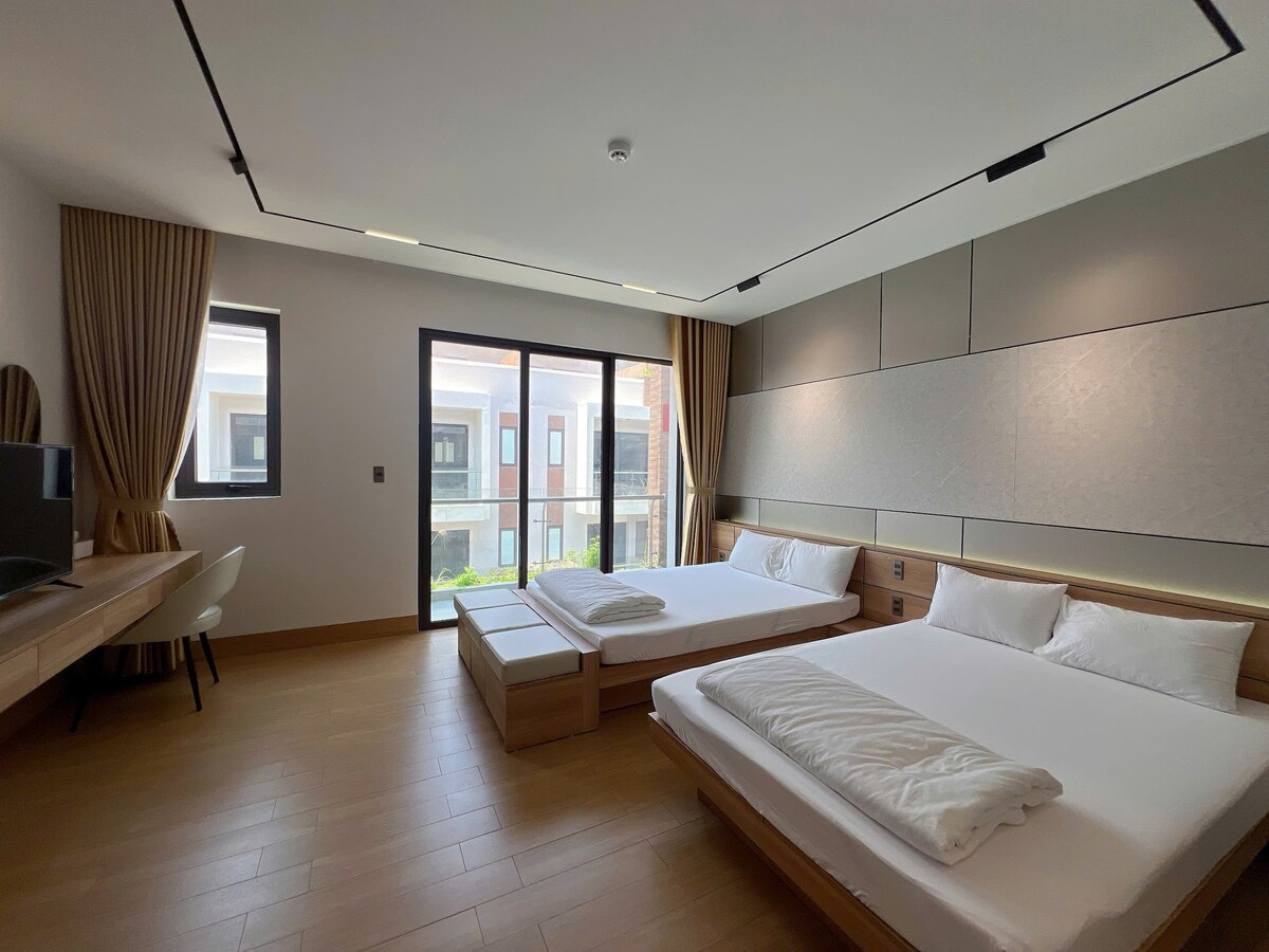 A spacious guest room features two beds and large windows allowing natural light to fill the space. A modern desk and chair are positioned near the window, and light wood finishes create a calming atmosphere. Free Wi-Fi and a view of the surrounding area are notable features.
