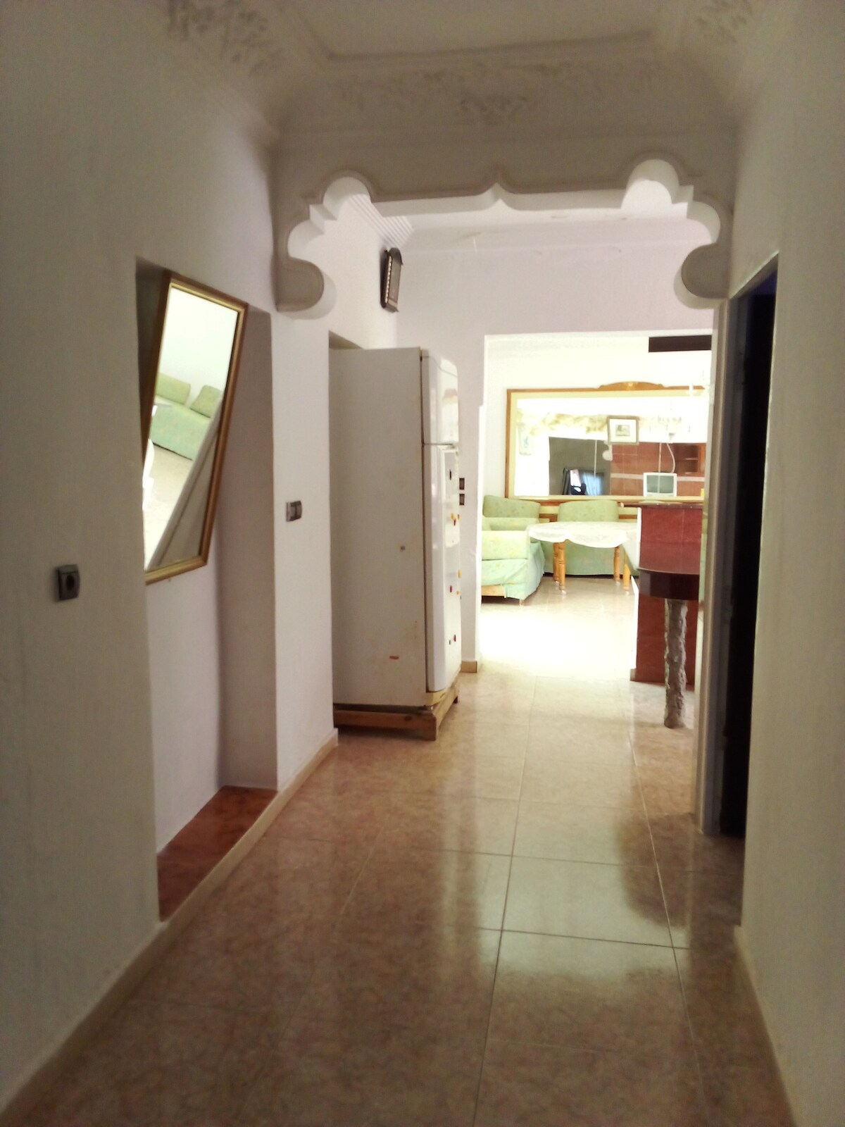 A well-lit corridor is visible, featuring smooth tiled flooring and walls in a light hue. A large mirror hangs on one side, reflecting the space. At the end of the corridor, a glimpse of a living area with green seating can be seen, creating an inviting transition.