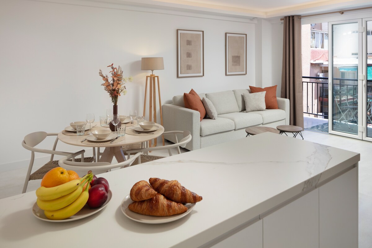 An open kitchen and living area is displayed, featuring a modern design. A round dining table is set for four, with tableware neatly arranged. A comfortable sofa with cushions is positioned nearby. Fresh fruit and croissants are presented on the kitchen countertop, enhancing the inviting ambiance.