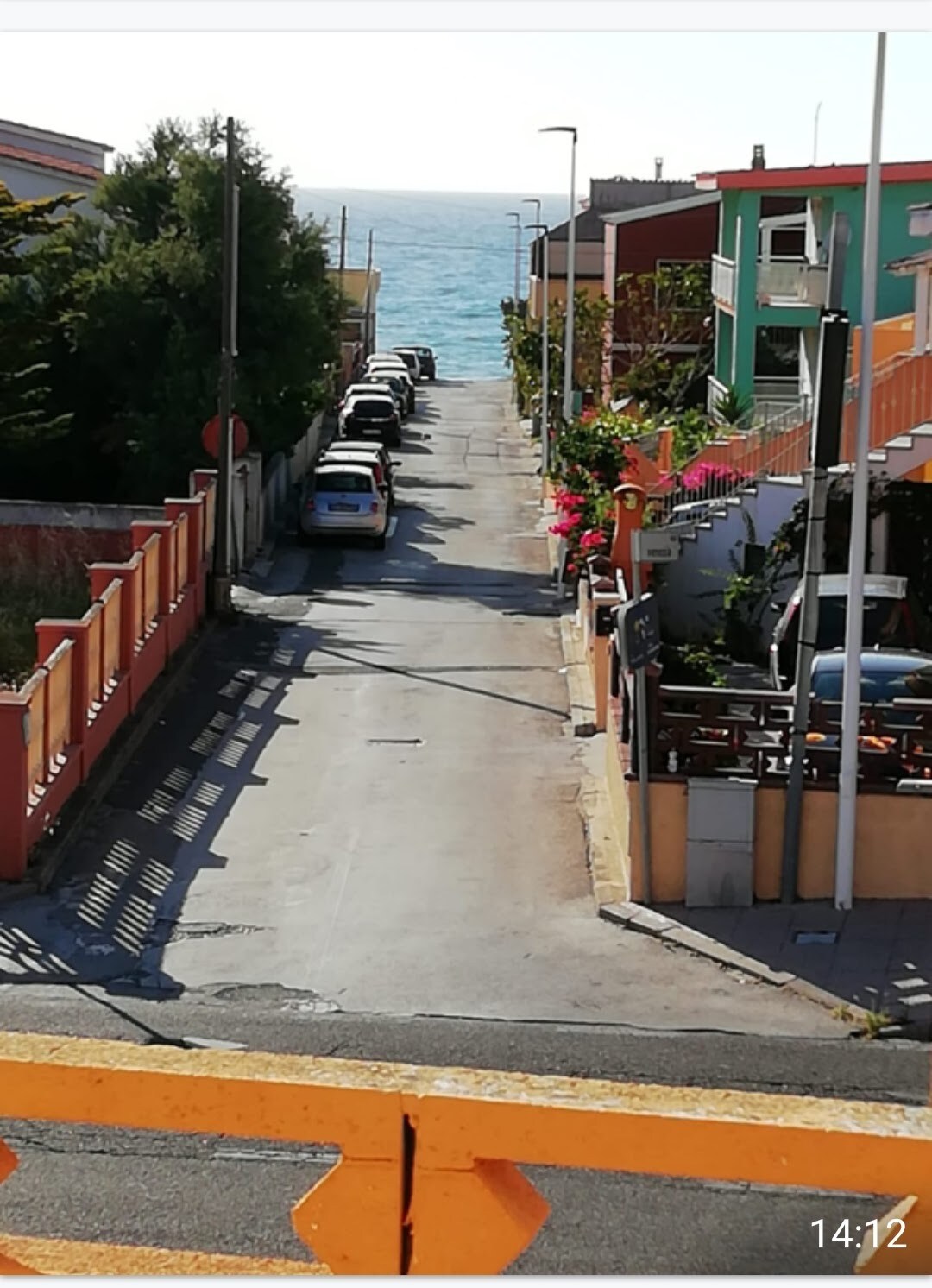 A narrow street lined with parked cars leads towards the sparkling blue sea in the background. Colorful buildings with vibrant flowers frame the street, creating a pleasant atmosphere. The sun brightens the scene, highlighting the peacefulness of the surroundings.