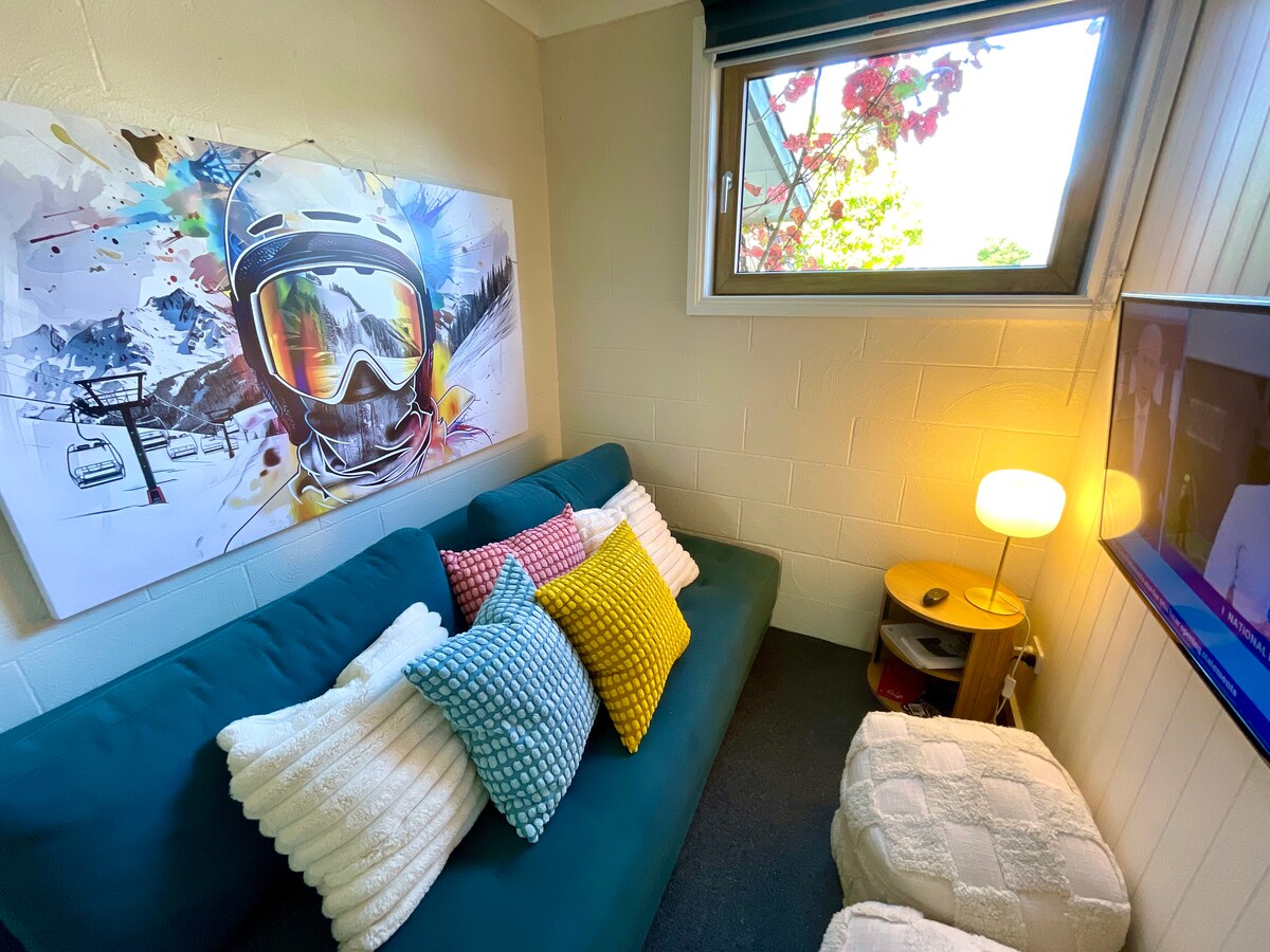 A cozy seating area features a teal sofa adorned with an assortment of colorful pillows. Sunlight filters through a nearby window, illuminating the space. A lamp provides soft lighting, and a small side table holds a decorative piece. A vibrant mountain-themed artwork adds visual interest.