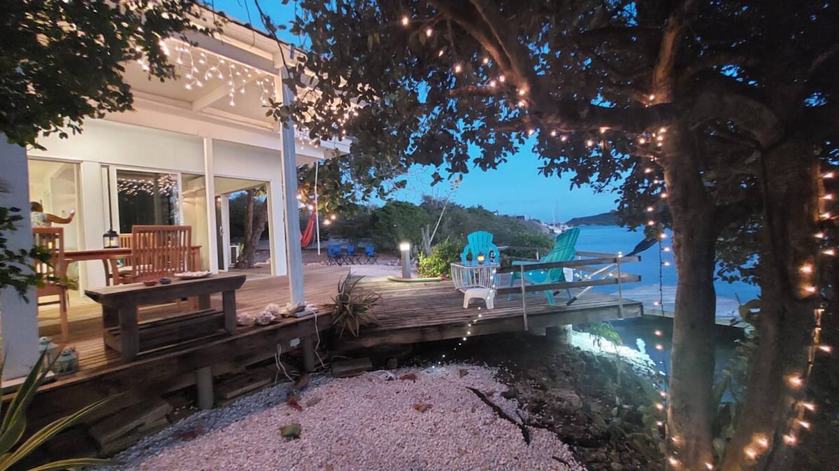 A serene outdoor area is illuminated by string lights, providing a cozy ambiance. Comfortable seating options, including a dining table and brightly colored chairs, are placed on a wooden deck that extends toward the water's edge, surrounded by natural greenery.