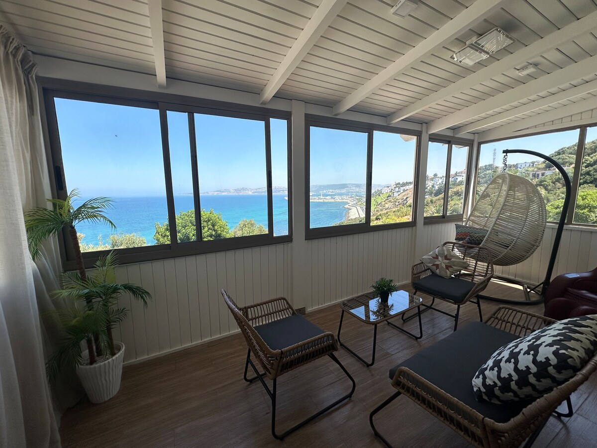An inviting sunroom features large windows that provide panoramic views of the ocean and coastline. Comfortable seating arrangements, including a hanging chair and lounge chairs, surround a small coffee table. Lush greenery is positioned in a planter, enhancing the serene environment.