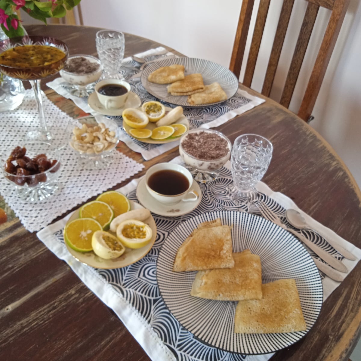 A wooden table is set for breakfast, featuring white plates with striped designs. Two cups of coffee, fresh fruit, baked pastries, and a dish of dates are arranged thoughtfully. Clear glasses add a touch of elegance, while a crocheted tablecloth complements the setting.