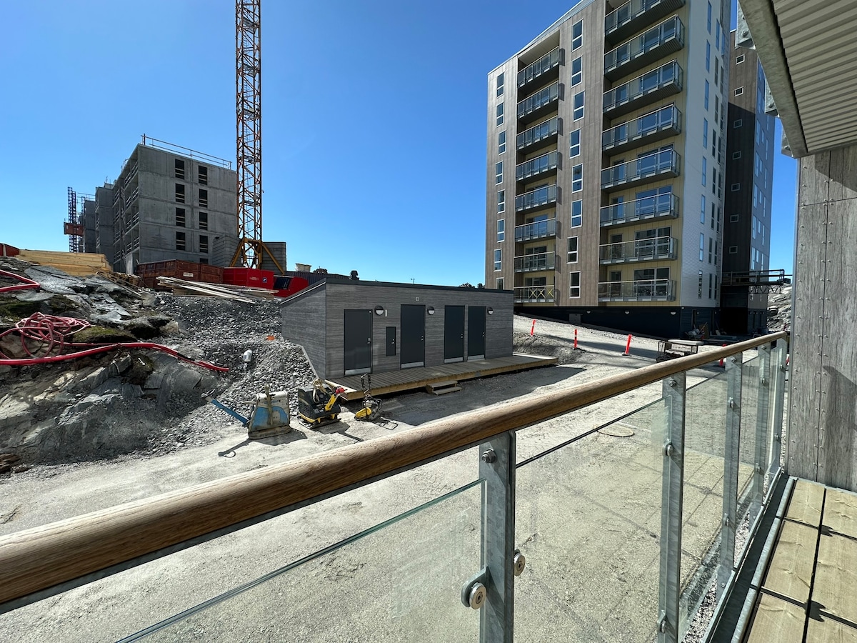 A balcony view is offered, featuring surrounding construction activities. The foreground shows a gravel surface with construction materials, while new buildings rise in the background amidst a clear blue sky. Glass railings frame the space, providing an open perspective of the area.