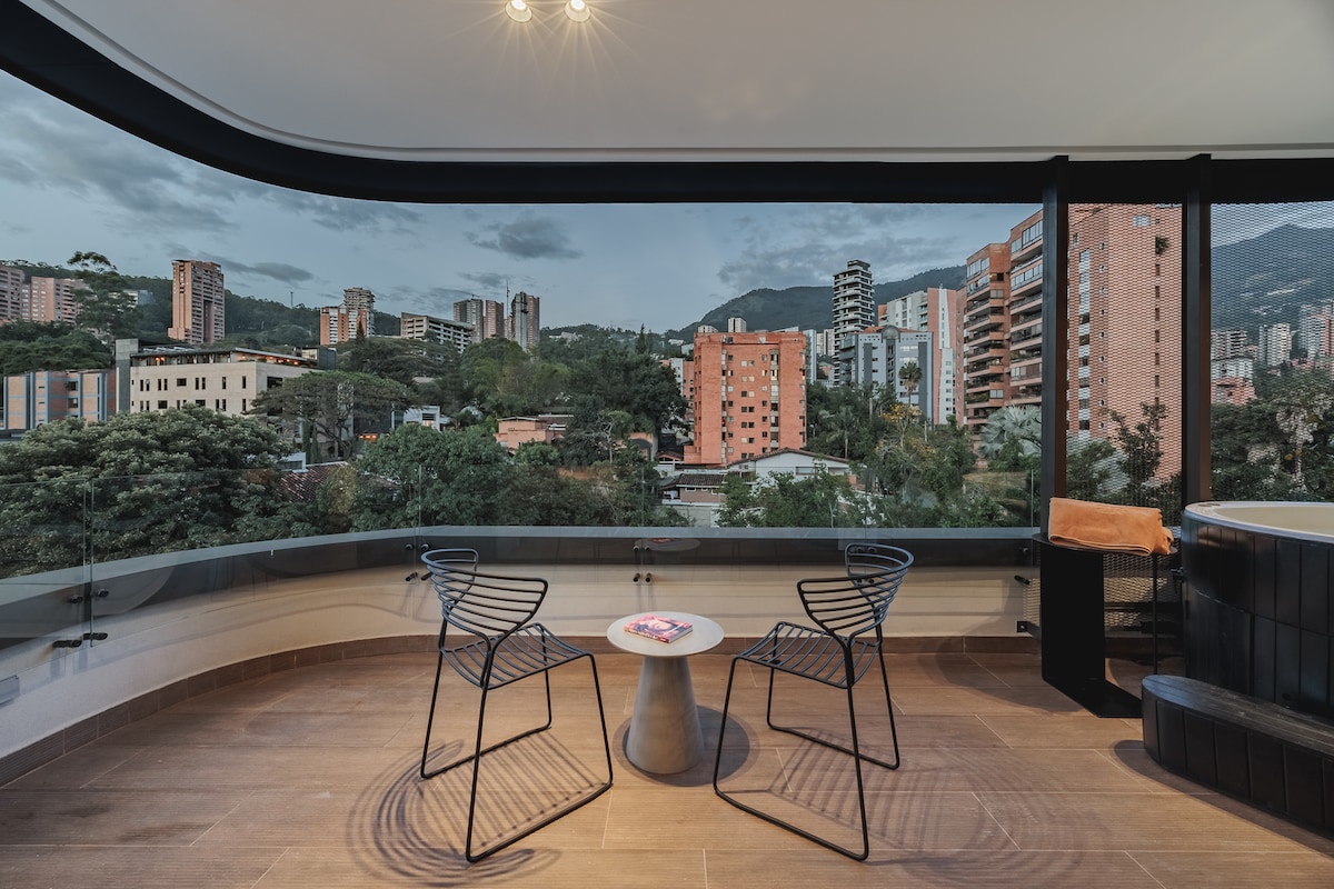 A modern balcony area features two sleek chairs positioned around a round table, offering a scenic view of the surrounding buildings and greenery. The backdrop includes a blend of urban architecture and natural landscapes, enhancing the spacious feel of the outdoor seating arrangement.