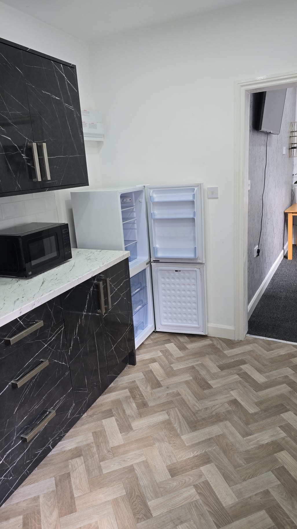 The kitchen features dark cabinetry with sleek marble countertops. A microwave is positioned on the counter, while a refrigerator with a freezer section stands in the corner. The herringbone-patterned floor adds a modern touch, and a doorway leads to an adjacent room.