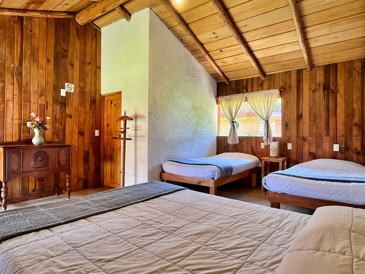 A cozy bedroom features wooden walls and a vaulted ceiling. A spacious double bed sits alongside three individual beds. Natural light enters through a window adorned with sheer curtains. A wooden cabinet adds character to the room, complementing the warm tones of the decor.