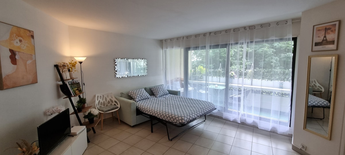 A comfortable living space is featured, showcasing a spacious bed with a patterned coverlet. Natural light floods the room through large glass doors that open to a lush terrace. A cozy chair and a television complete the area, with artwork adorning the walls.