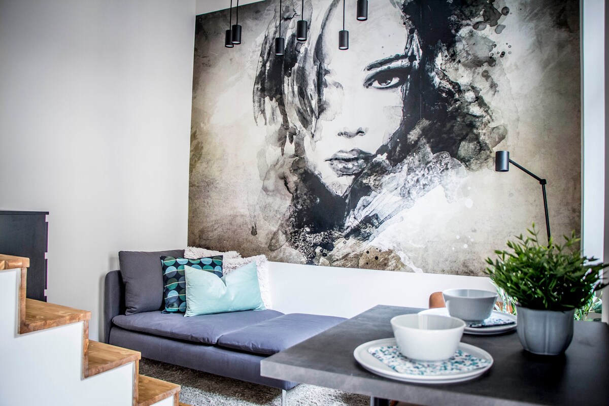 A modern living area features a large wall mural of a woman, adding an artistic flair. A sleek gray sofa is adorned with blue and white cushions. A dining table is set with two bowls, complemented by a small plant on the table.