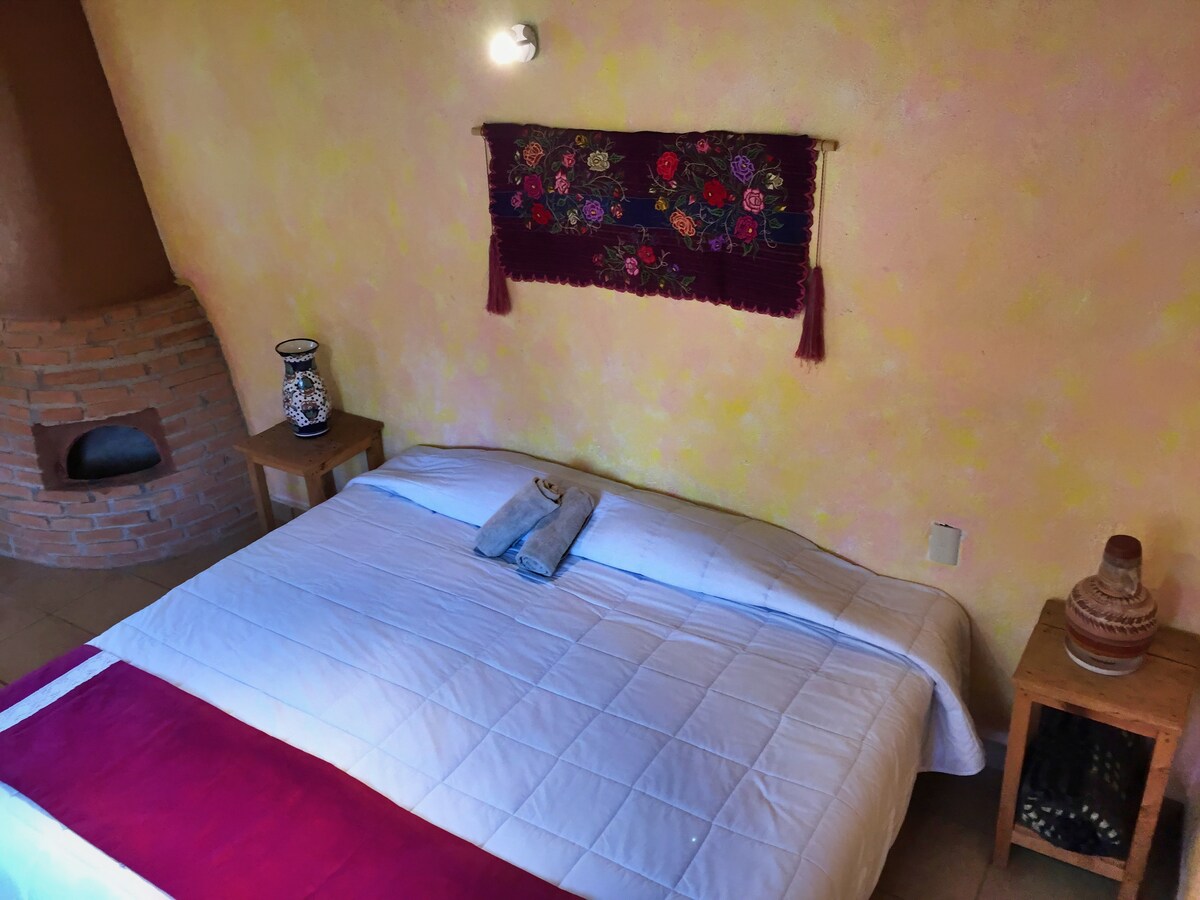 A spacious bed is dressed in white bedding with a decorative red accent. Two towels are neatly arranged on the bed. A rustic brick fireplace is visible on the left, and colorful textile art hangs on the wall above the headboard.