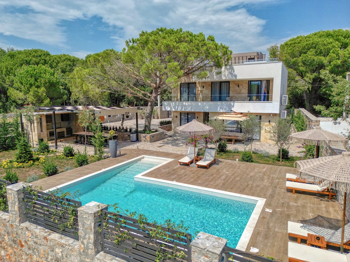 A modern villa is surrounded by lush greenery, featuring a spacious pool area with sun loungers and shaded umbrellas. The outdoor space includes a wooden deck and mature trees, providing a peaceful setting. The villa's facade showcases large windows and a contemporary design.