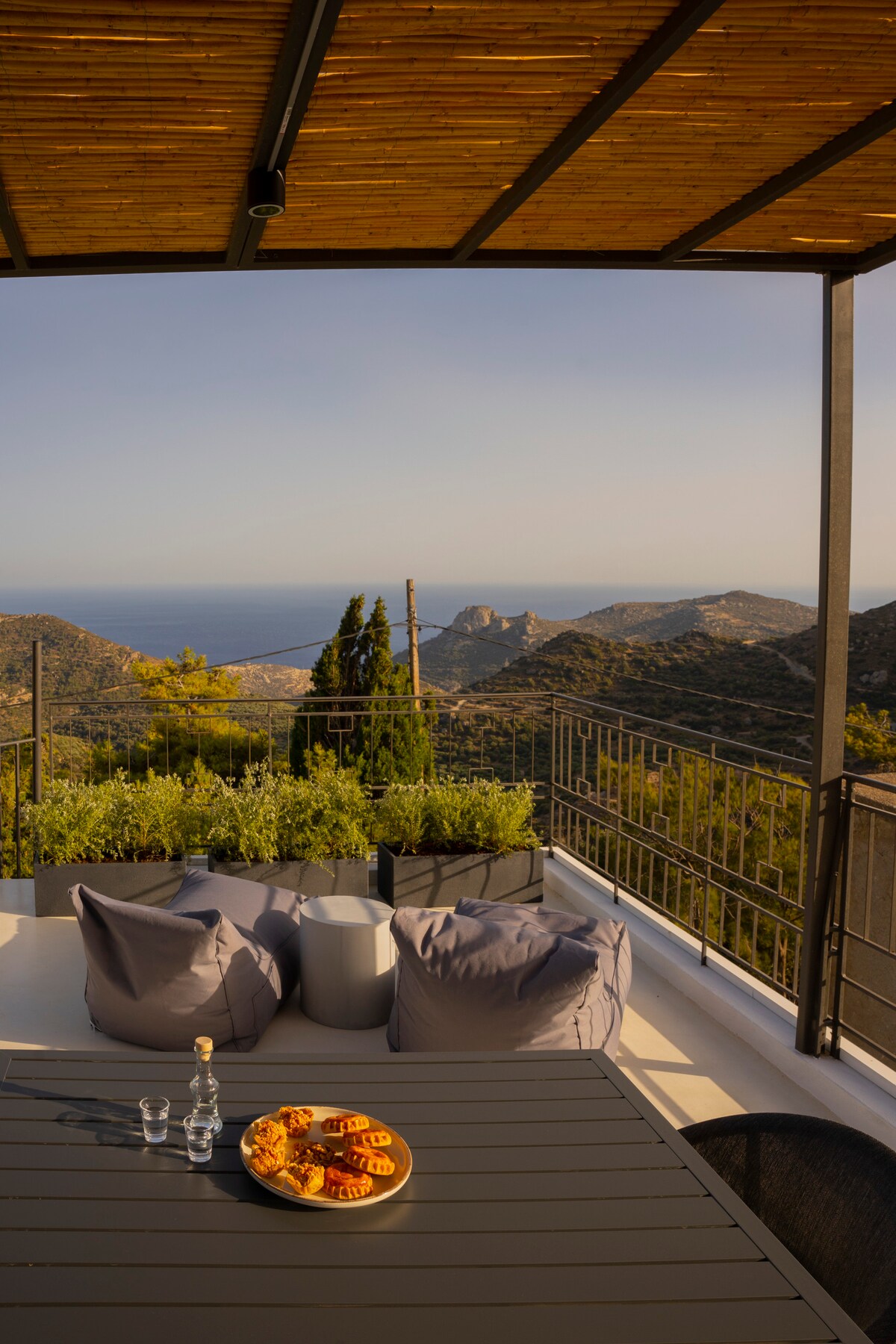 A spacious outdoor terrace features a dining table surrounded by comfortable chairs. Potted greenery adds a touch of nature, while a scenic view of the mountain and sea can be seen in the background. A plate of pastries and two bottles of water are placed on the table.