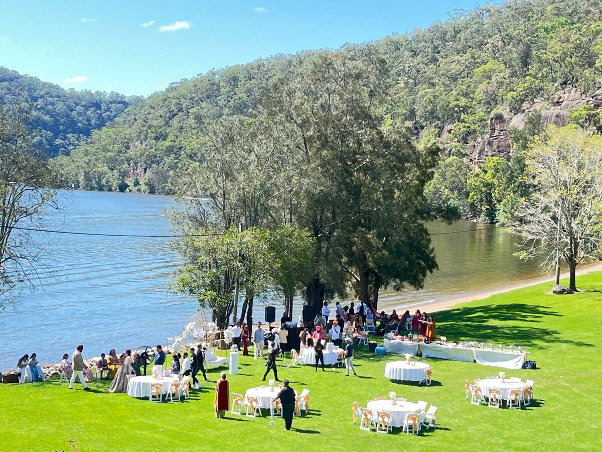 A scenic riverside gathering is depicted with a large grassy area dotted with white tables and chairs. Guests are mingling near the water's edge, surrounded by lush greenery and the tranquil backdrop of the river and hills.