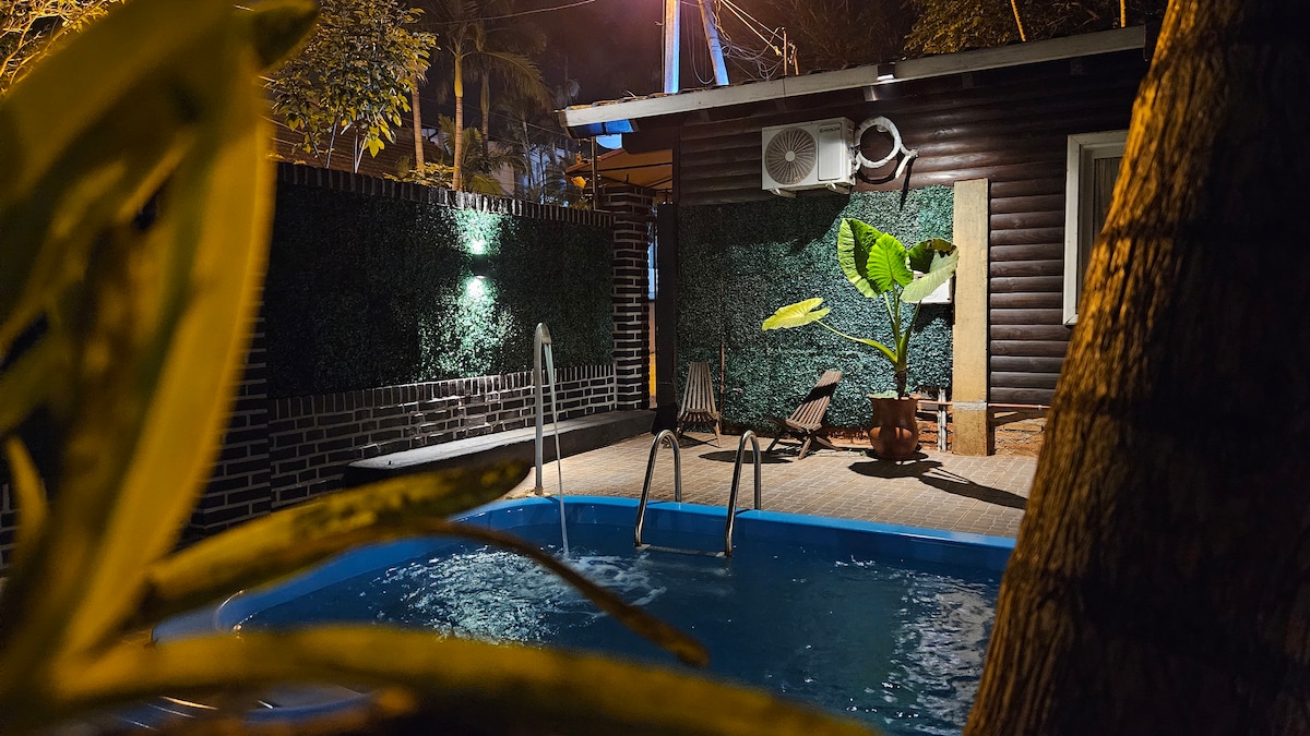 A refreshing pool is framed by greenery, featuring a ladder for easy access. The pool area is softly illuminated by wall-mounted lights, complemented by a large potted plant that adds a natural touch. The surrounding space includes lounge chairs for relaxation.