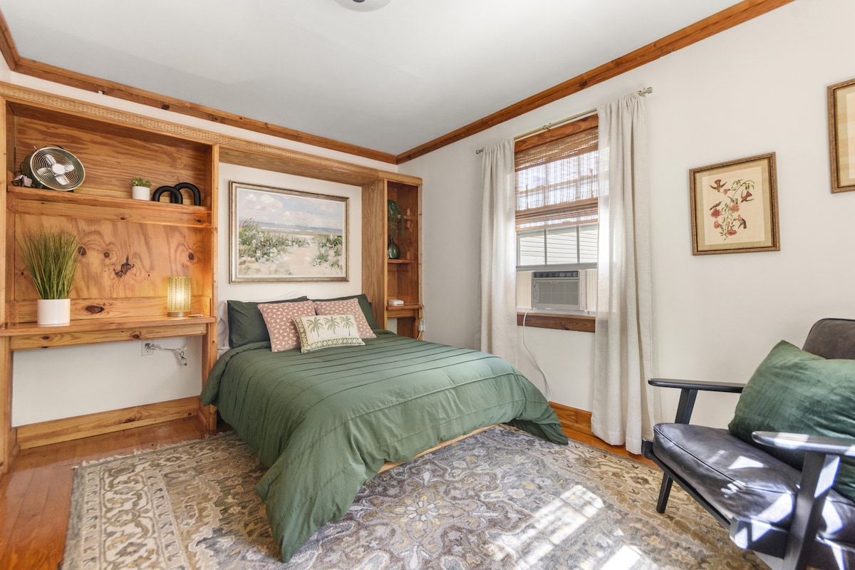 A cozy sleeping area features a spacious bed with a green coverlet and decorative pillows. Wooden shelving lines the wall, displaying a plant and decorative items. natural light streams in through the window, creating a bright and welcoming ambiance.