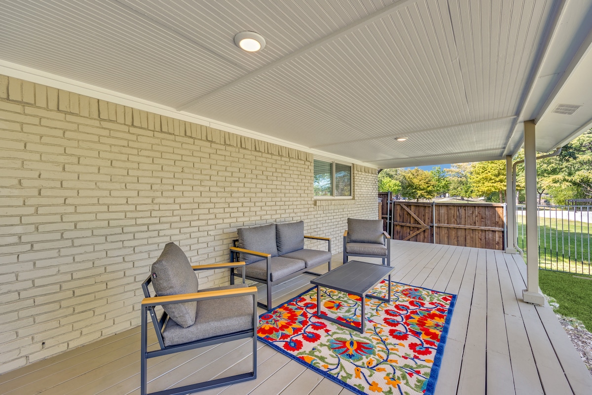 A covered patio area features a vibrant area rug with floral patterns at its center. Four patio chairs with cushioned seats surround a small rectangular table, providing a comfortable space for outdoor relaxation. Green grass is visible beyond the patio, with a wooden fence in the background.