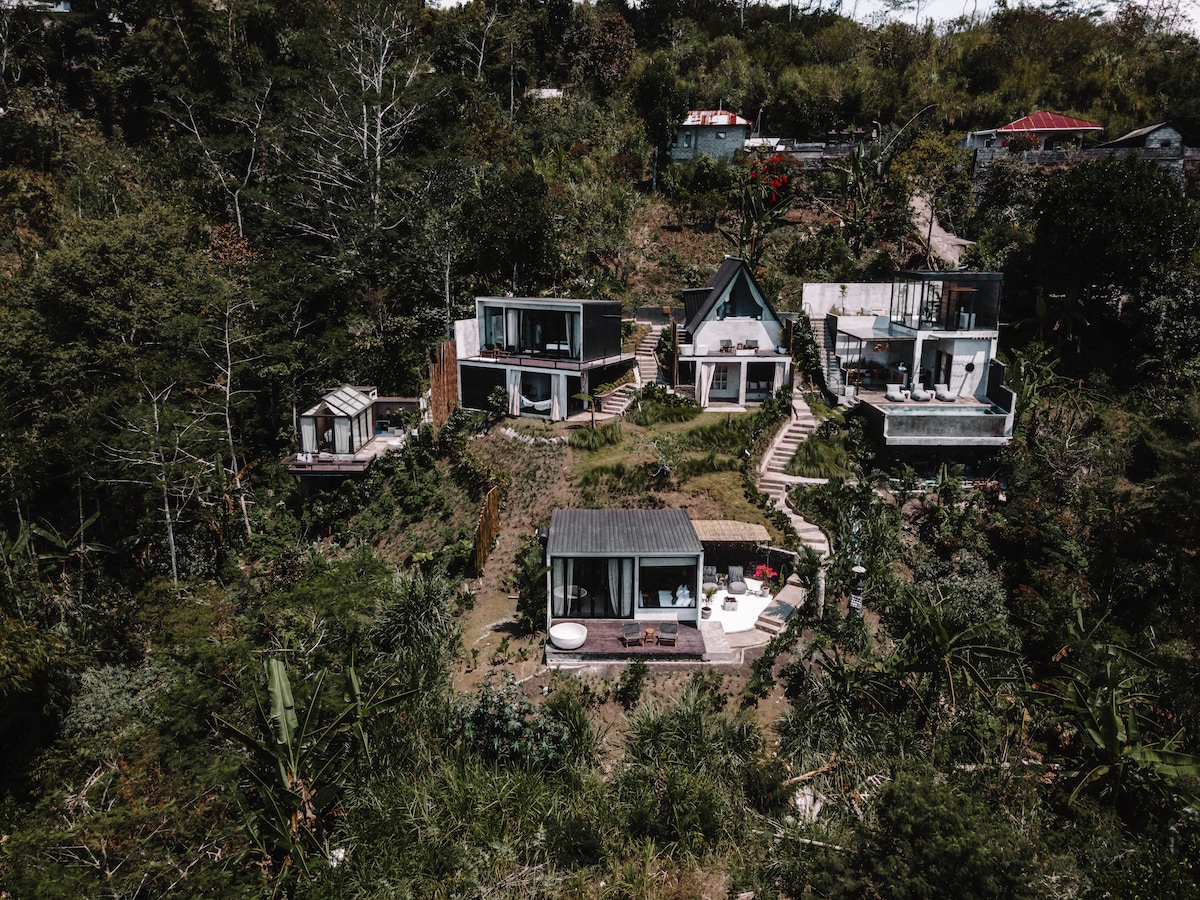 Aerial view of Batur Cabins situated on a hillside, surrounded by lush greenery. The layout features several cabins with distinct architectural styles, interconnected by winding pathways. A spacious outdoor area includes a terrace and an outdoor bathtub, offering a serene retreat within the natural landscape.