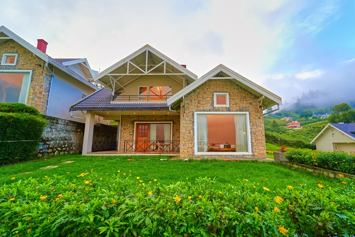 The villa showcases a charming stone exterior with large glass windows, surrounded by neatly trimmed green lawns and vibrant flowers. A spacious porch is visible, featuring a seating arrangement, while the roof is adorned with a distinctive sloping design. The tranquil landscape enhances the home's inviting presence.