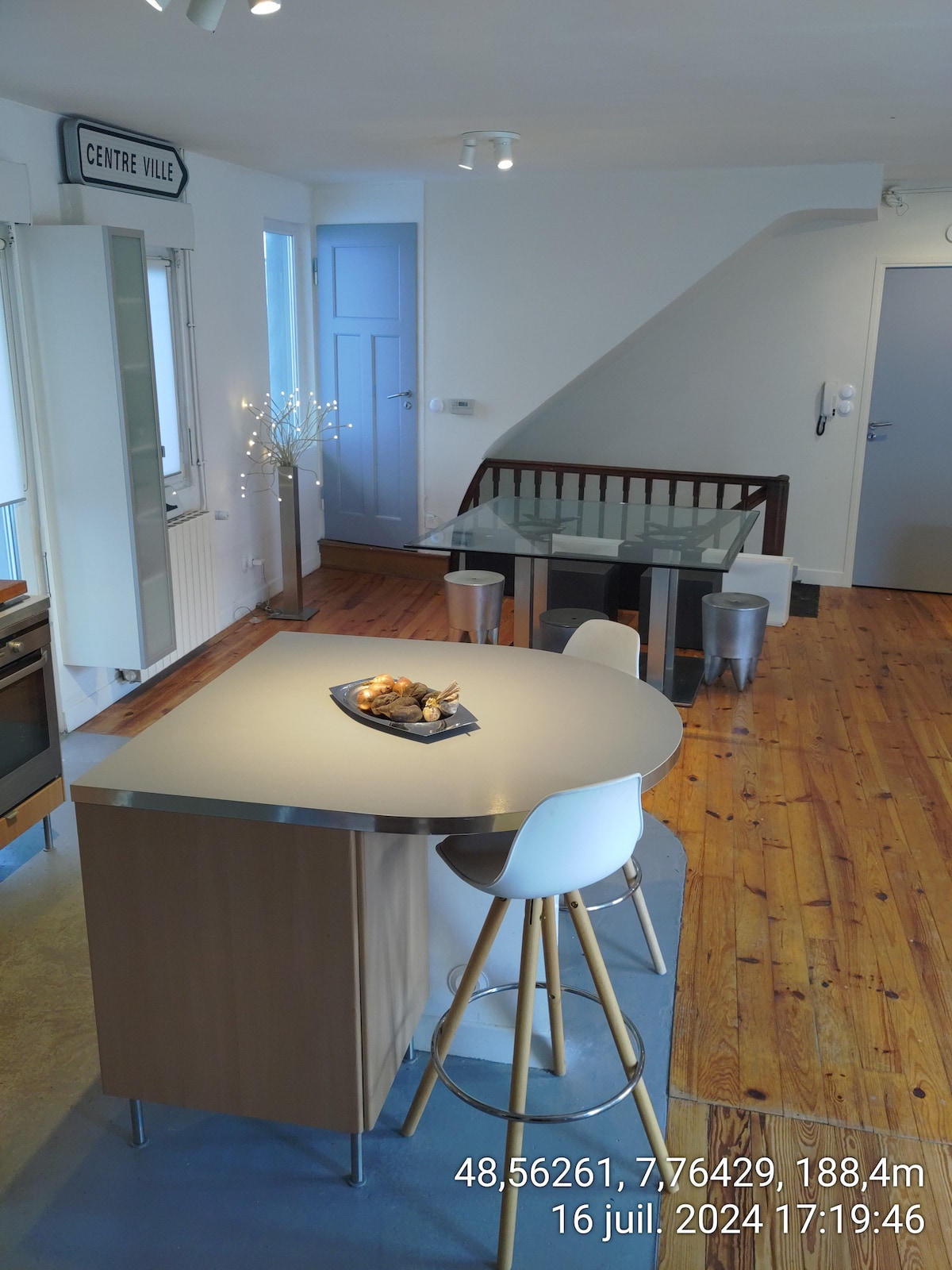 An open kitchen and dining area is shown, featuring a sleek kitchen island with a metallic countertop. A glass dining table is surrounded by modern stools. Light wood flooring adds warmth to the space, complemented by natural light entering through the windows.