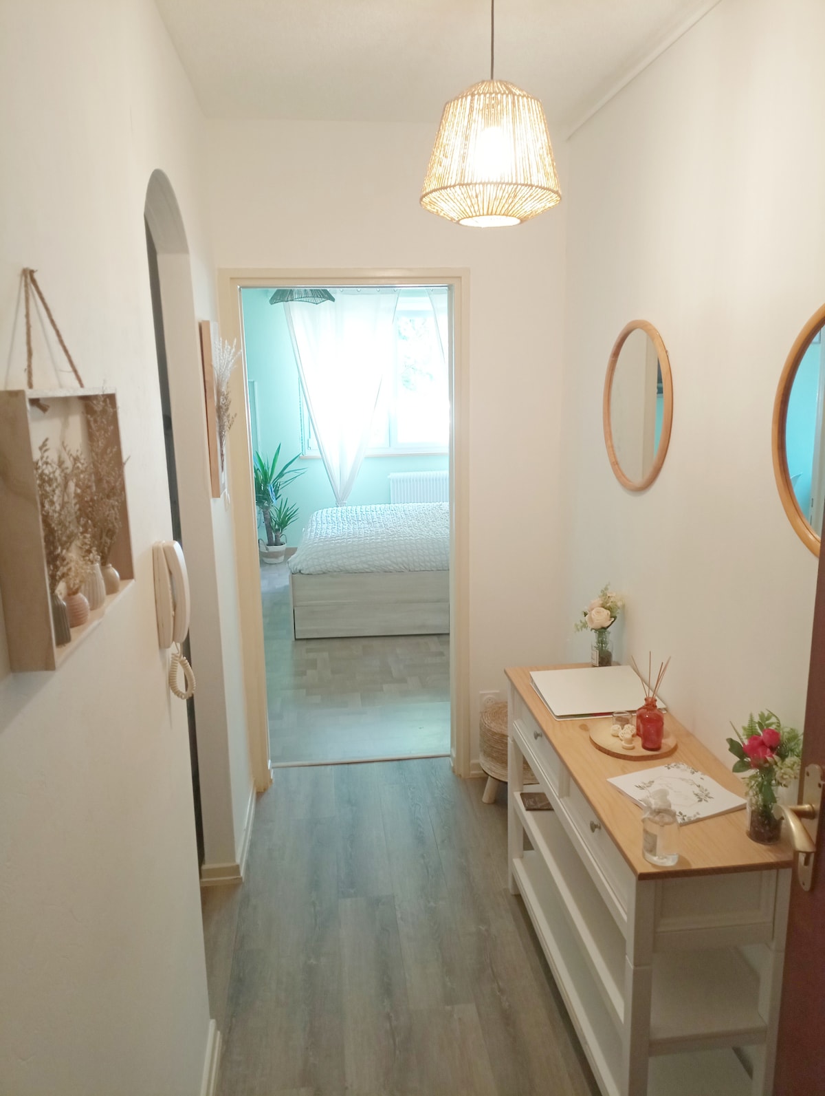 A well-lit hallway features light-colored walls and wooden flooring. A small console table sits against one side, adorned with a floral arrangement and decorative items. Two circular mirrors provide visual interest, while a glimpse of the bedroom is visible at the end of the corridor.