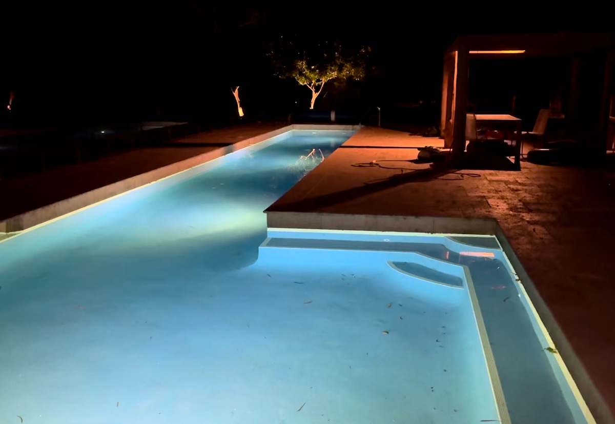 The lap pool is softly illuminated at night, casting a gentle blue glow over the water. Surrounding lounge areas are visible, with trees in the distance and highlights of nearby outdoor seating, creating a serene atmosphere for evening relaxation.
