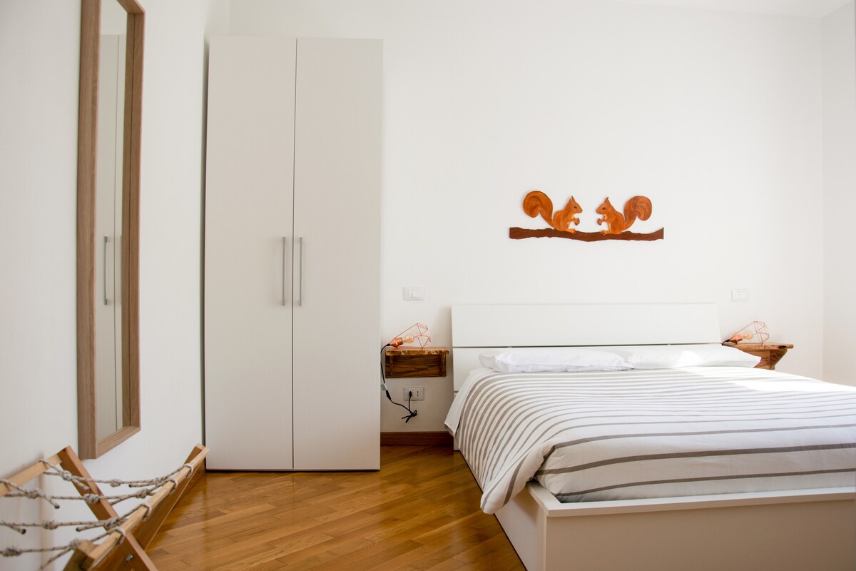 A simple, well-lit bedroom features a spacious bed with striped linens. A wooden headboard with squirrel motifs adds a charming touch above the bed. An elegant mirror and a wardrobe are positioned against the white walls, complemented by a wooden floor that enhances the room's warmth.