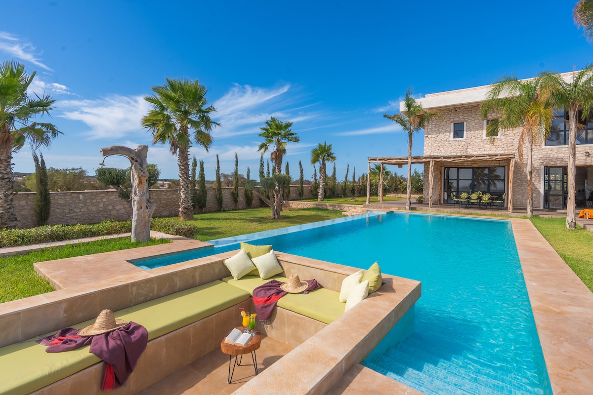 A spacious patio area surrounds a glistening pool, featuring a sunken seating area adorned with colorful cushions. Palm trees frame the scene, complemented by a stone building in the background. The clear blue sky enhances the inviting atmosphere of the outdoor space.