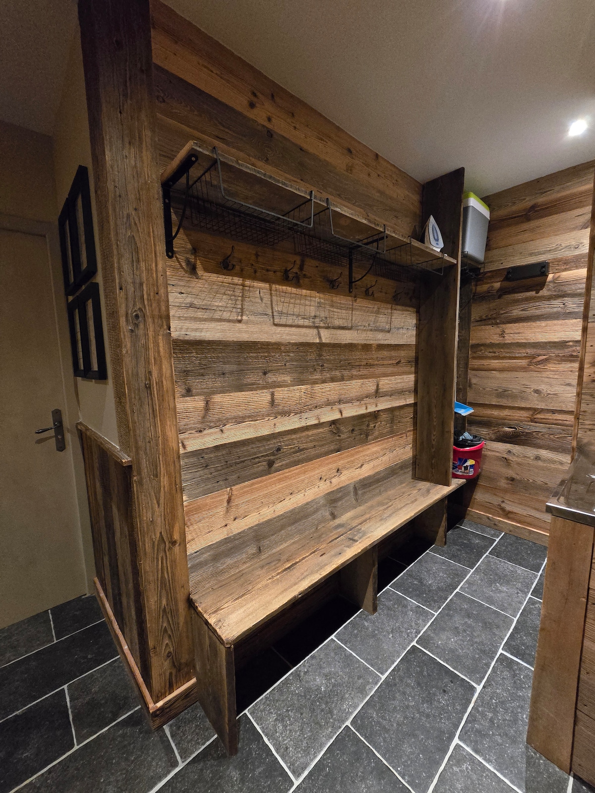A functional entryway features a built-in wooden bench with slatted seating and a wall with rustic wooden panels. Above the bench, a metal rack provides space for hanging items. The floor is laid with dark slate tiles, complementing the warm wood tones.