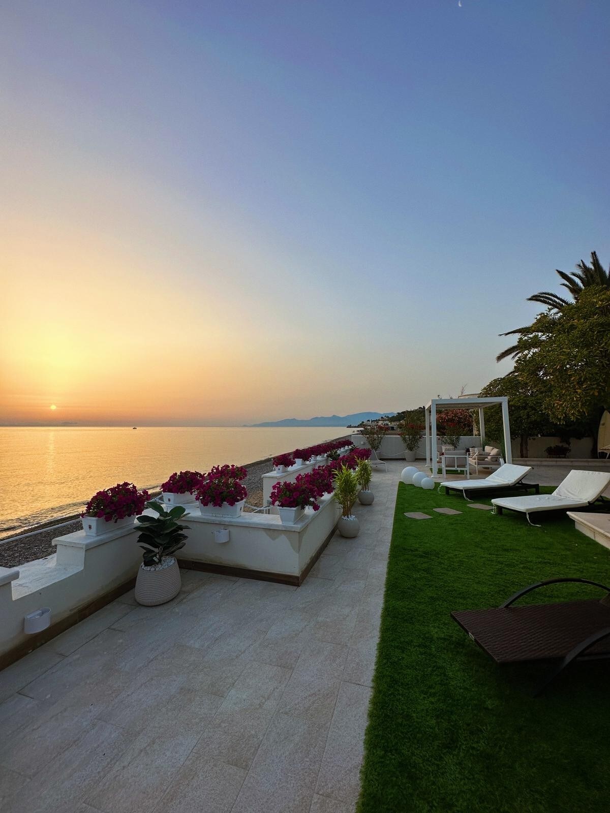 A serene terrace overlooks a calm sea during sunset, filled with pink and purple flowers along the railing. Sun loungers are positioned for relaxation, with a small patch of green grass adding a touch of nature to the space. The distant silhouette of mountains completes the tranquil view.