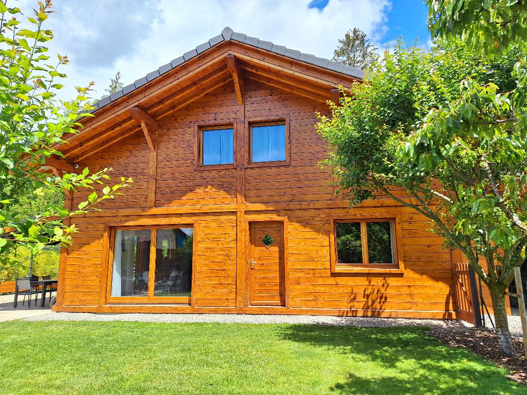 The chalet features a warm wooden exterior, characterized by a gabled roof and large windows. A well-maintained lawn surrounds the building, and trees provide natural greenery. The front door is accentuated by a small diamond-shaped window.