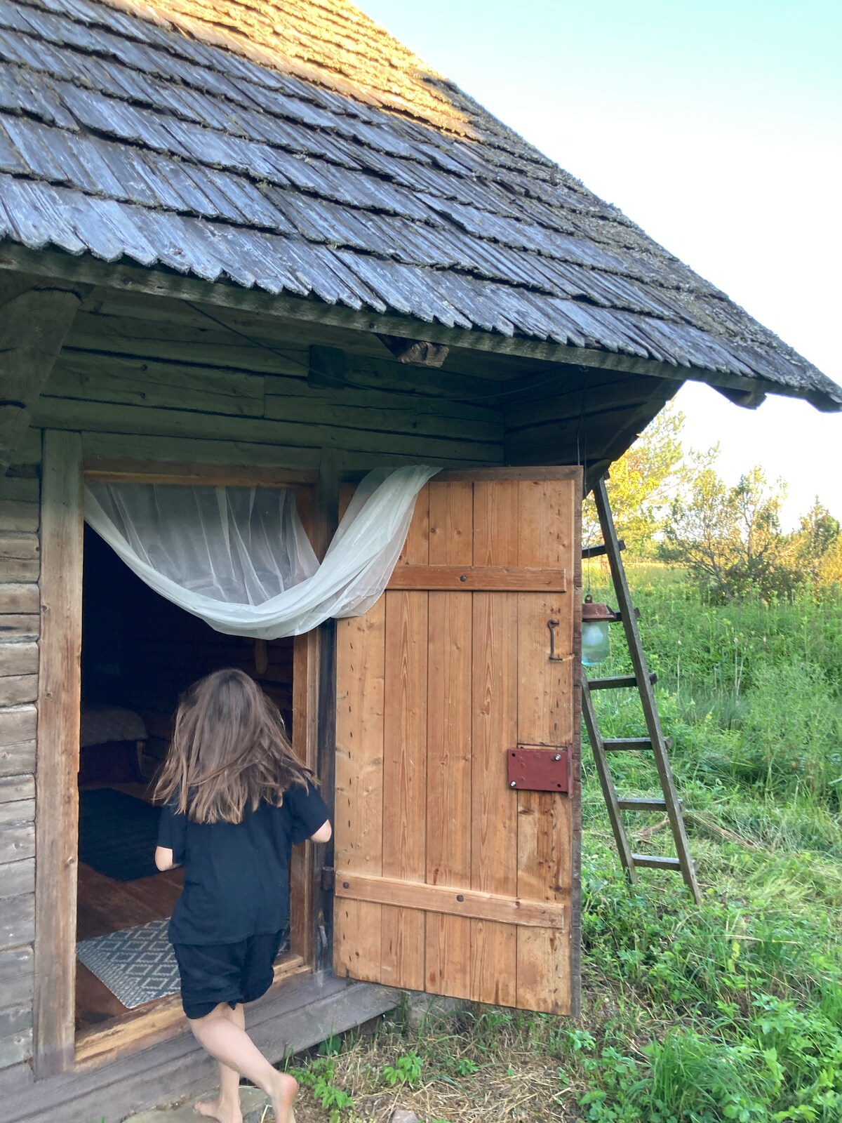 A traditional wooden door of the barn room is open, revealing a cozy interior. A sheer curtain gently sways at the entrance. Lush greenery surrounds the building, and a wooden ladder leans against the structure, inviting exploration of the countryside.