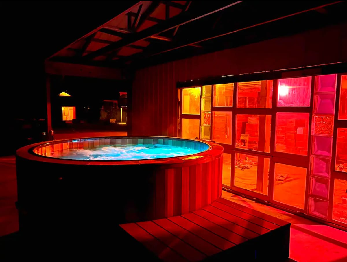 A large cedar hot tub is illuminated with soft blue light, creating a calming atmosphere. The surrounding area is highlighted by colorful lighting from nearby structures, enhancing the outdoor setting at night.