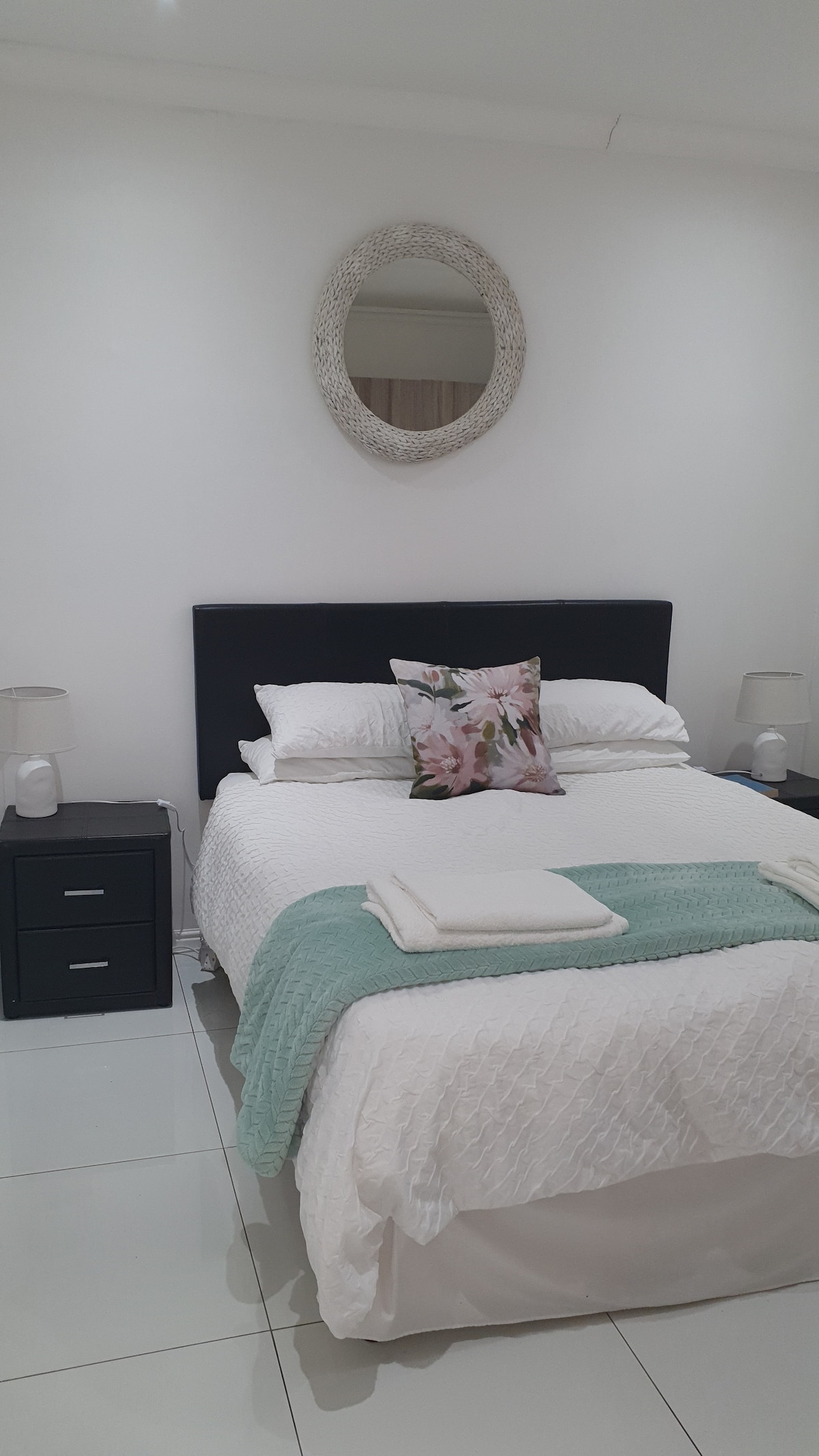 The bedroom features a spacious bed centered against a white wall. Flanking the bed are two nightstands with lamps. A decorative mirror hangs above the bed, while soft linens and a light turquoise throw blanket adorn the bed, adding a touch of color.