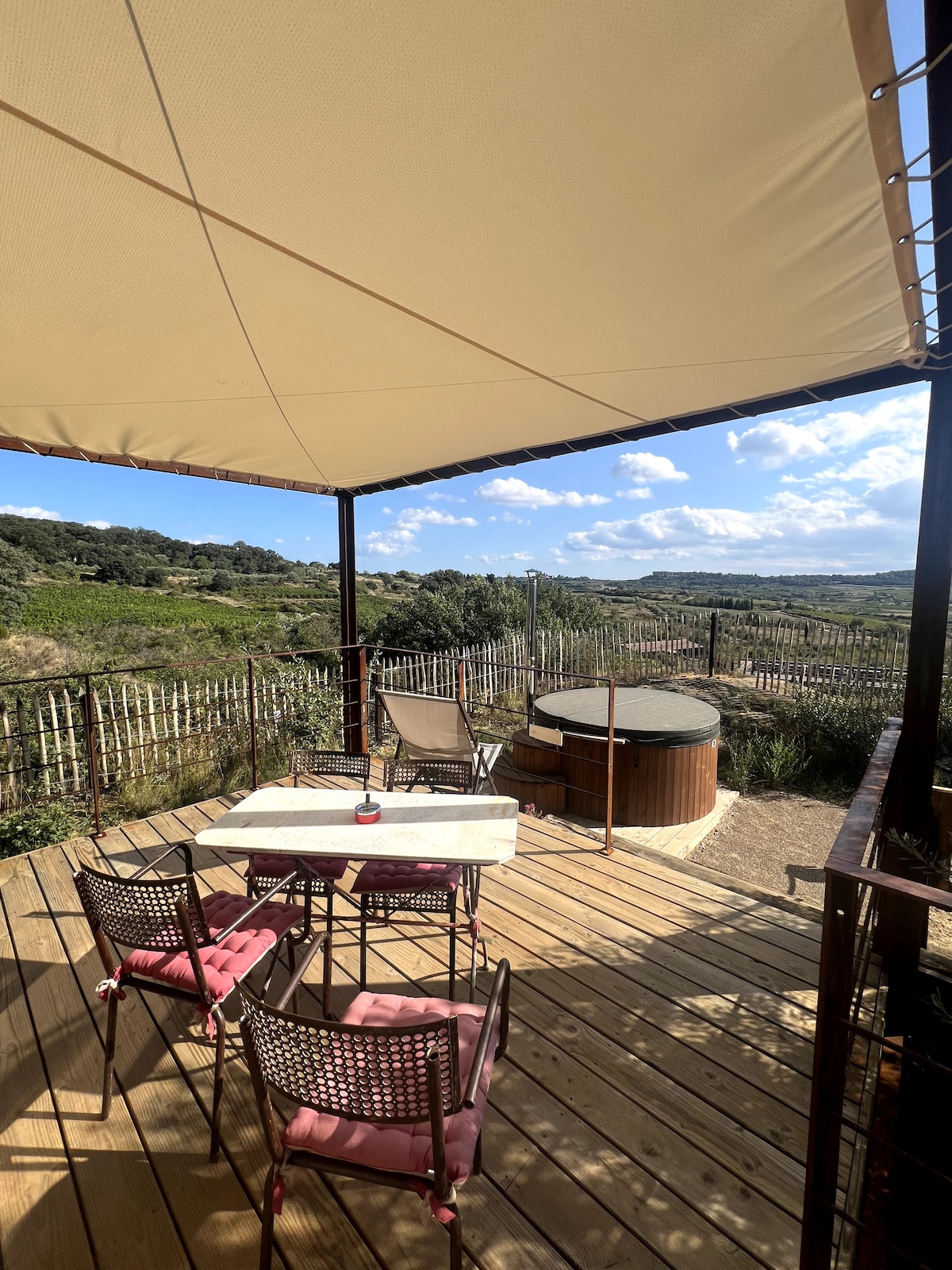 A private terrace is shown, featuring a wooden table with seating for four. A circular wooden Swedish bath is positioned nearby, surrounded by greenery. The space is sheltered by a large canopy, allowing for outdoor enjoyment while protecting from direct sunlight.
