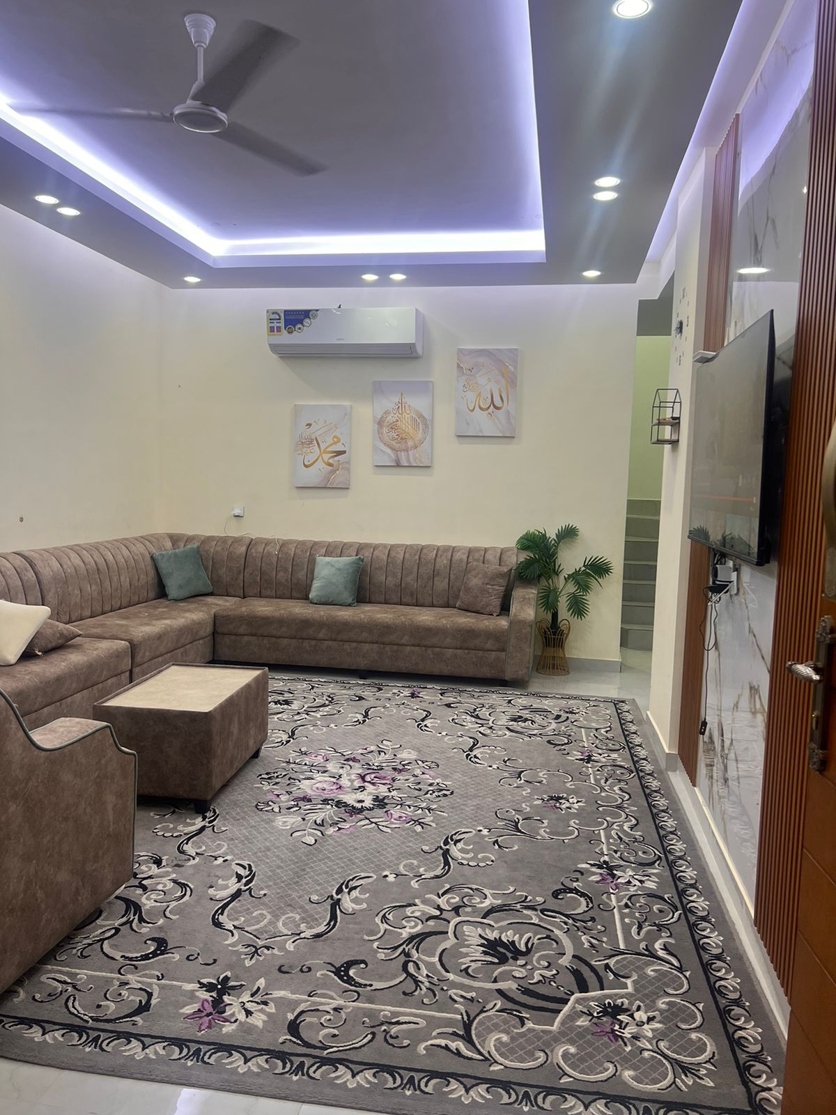 A spacious living area is furnished with a sectional couch and a comfortable armchair. A large rug with intricate floral patterns anchors the space. Bright artwork adorns the walls, and modern lighting creates a warm ambiance, complemented by a small plant adding a touch of nature.