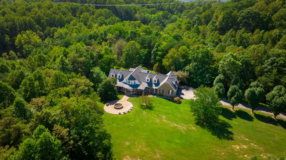Aerial view of a spacious private estate surrounded by lush greenery and trees, featuring a large landscaped yard. The multi-level home is complemented by a circular fire pit area, offering an inviting space for outdoor gatherings.