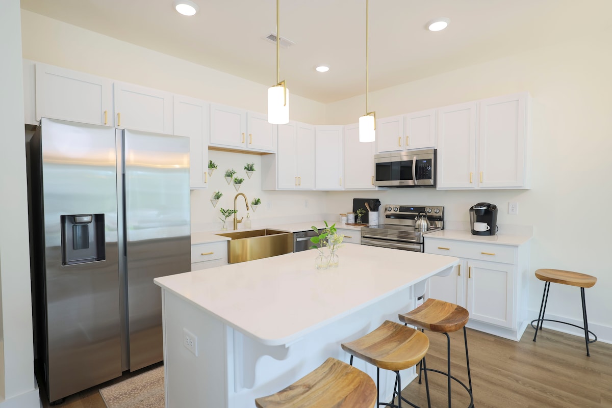 The modern kitchen features stainless steel appliances, including a refrigerator and microwave. A golden sink complements the white cabinetry, while an island with seating for three adds functionality. Soft lighting illuminates the space, highlighting the greenery on the wall that enhances the fresh ambiance.