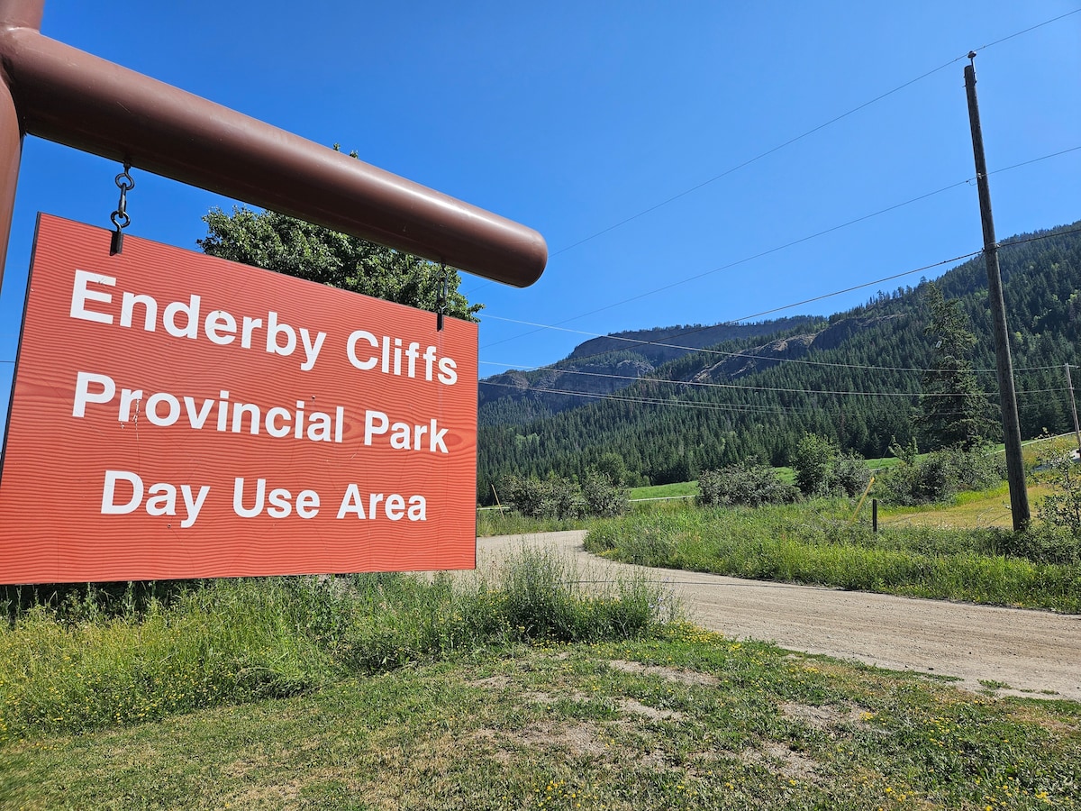 A red sign displaying 'Enderby Cliffs Provincial Park Day Use Area' is prominently featured. The sign is positioned near a dirt road leading into natural scenery filled with trees and rolling hills under a clear blue sky.