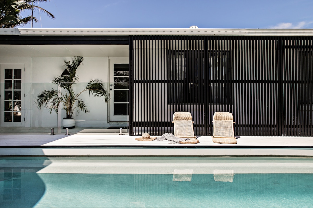 A modern outdoor area is displayed, featuring a shimmering pool in front of a striking black slatted screen. Two woven lounge chairs are positioned at the water's edge, accompanied by a round, natural fiber beach mat and a soft gray towel.