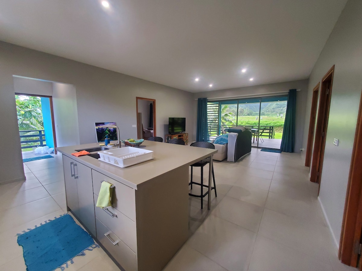 An open-concept living space features a modern kitchen island with stools, leading into a lounge area with a comfortable sofa and a television. Large sliding doors provide views of the lush surroundings and allow natural light to fill the room.