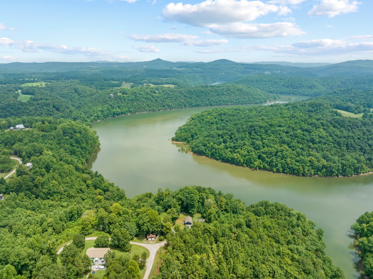 Aerial view captures the winding curves of Dale Hollow Lake, surrounded by lush greenery. The landscape features rolling hills that blend seamlessly with the water, showcasing a serene and tranquil environment. The sky reflects soft blue hues with scattered clouds.